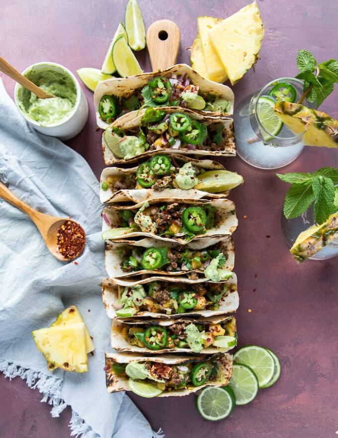 Lamb tacos on a taco holder surrounded by avocado sauce, pineapple salsa. lime wedges