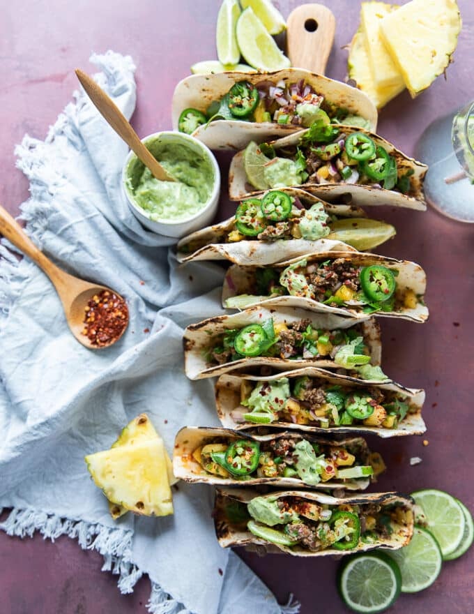 Finished Lamb Tacos assembled on a taco holder surrounded by pineapple salsa, crema and lime wedges, jalapeno slices