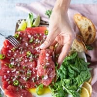 A hand holding a tuna carpaccio served over crunchy bread like a crostini