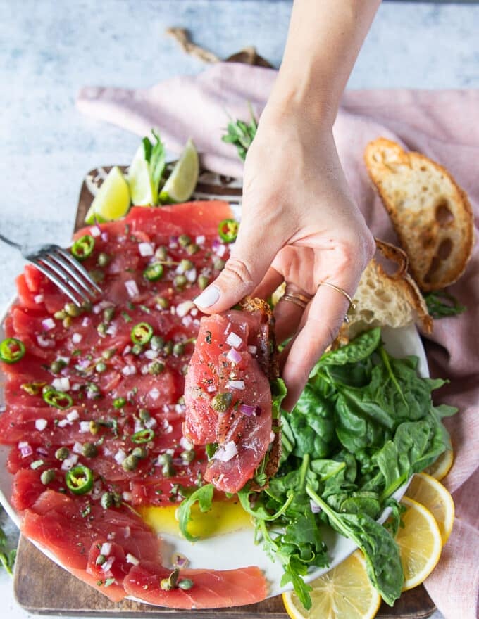 A hand holding a tuna carpaccio served over crunchy bread like a crostini