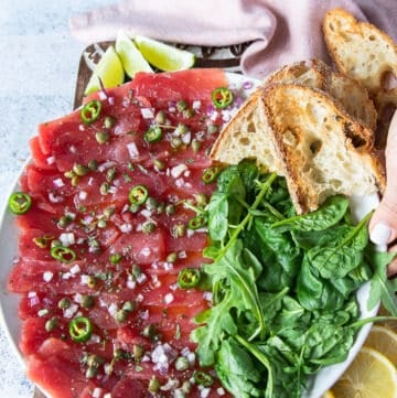 A round plate of tuna carpaccio with a side of arugula and crusty baguettes, lime wedges, and drizzled with capers, lime, scallions and olive oil vinaigrette