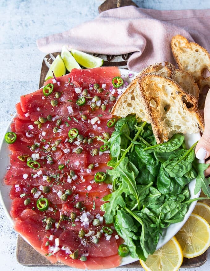 A round plate of tuna carpaccio with a side of arugula and crusty baguettes, lime wedges, and drizzled with capers, lime, scallions and olive oil vinaigrette