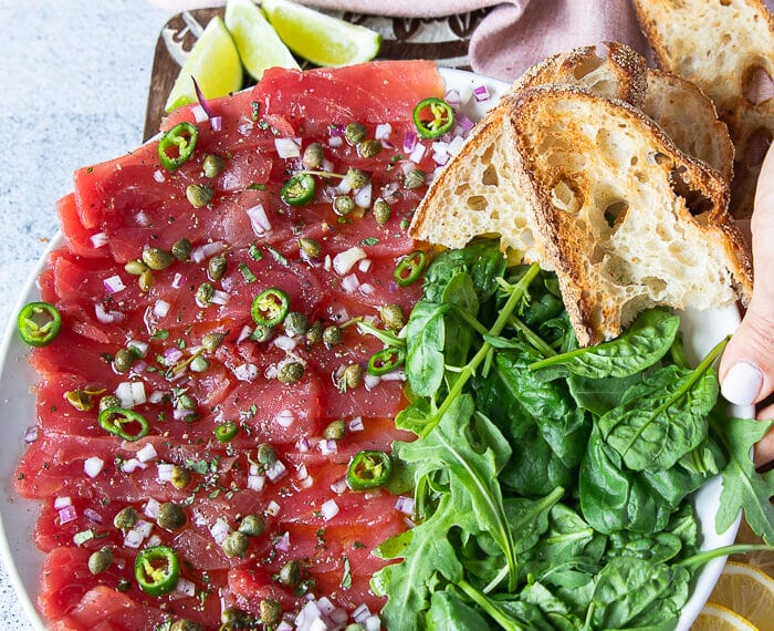 A round plate of tuna carpaccio with a side of arugula and crusty baguettes, lime wedges, and drizzled with capers, lime, scallions and olive oil vinaigrette