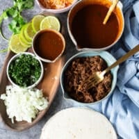 Ingredients to make birria quesadillas including: tortillas, birria meat, birria stew, cheese, cilantro, white onions chopped, jalapenos