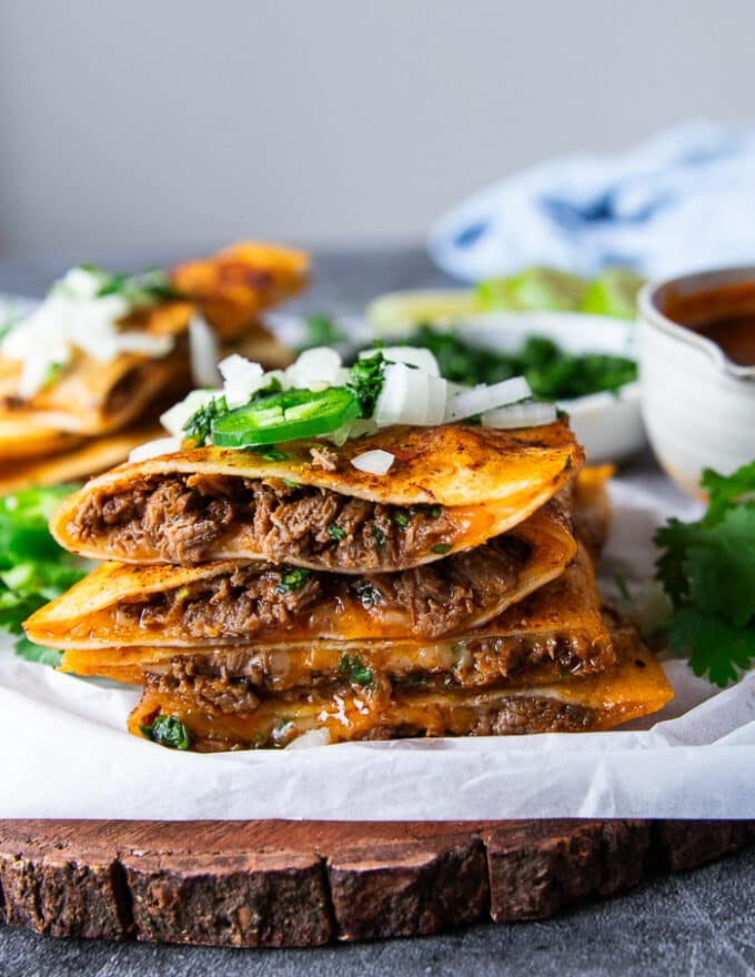 Cut up and stacked Birria quesadillas on a wooden board showing the birria meat and cheese filling and sauce on the tortilla. Topped with onions, cilantro and jalapenos