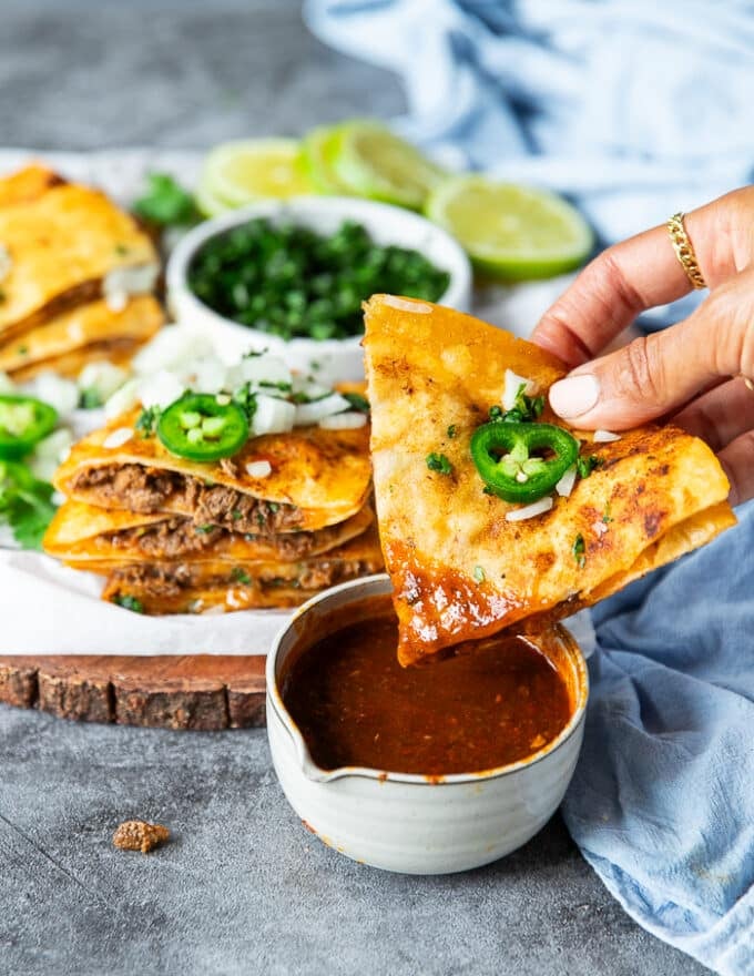 A hand dipping the birria quesadilla right into the bowl of birria stew