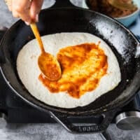 A hand slathering the tortillas with the birria stew to cover it and flavor it really well on the pan