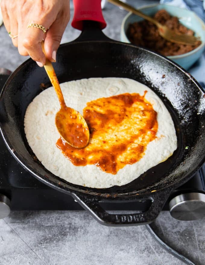 A hand slathering the tortillas with the birria stew to cover it and flavor it really well on the pan