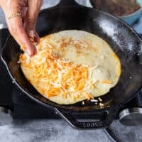 A hand scattering some shredded cheese al over the tortillas to cover it (while still int he pan)