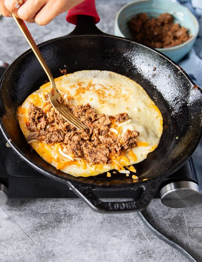 A fork adding a generous amount of birria meat over the cheese on the tortilla