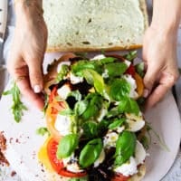 Basil leaves are added over the cheese in the caprese sandwich and a hand drizzling some balsamic syrup