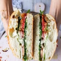 A hand holding a cut up caprese sandwich showing all the layers of basil mayo, fresh basil, heirloom tomatoes, mozzarella and some burrata cheese with balsamic syrup