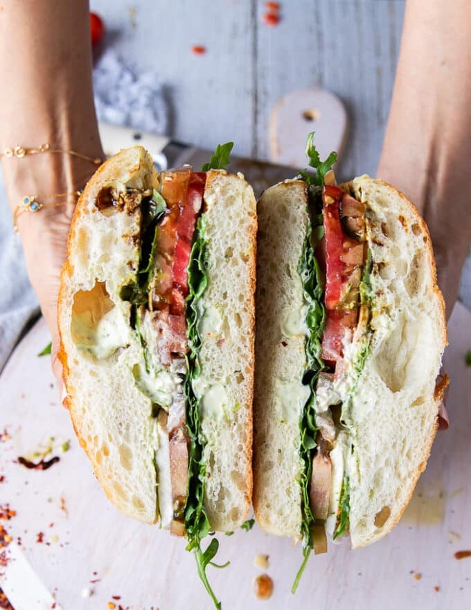 A hand holding a cut up caprese sandwich showing all the layers of basil mayo, fresh basil, heirloom tomatoes, mozzarella and some burrata cheese with balsamic syrup