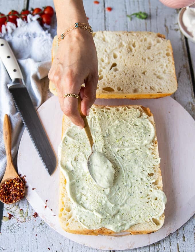A hand spreading a layer of basil mayo over the bread to start making caprese sandwich 