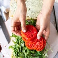 A hand assembling the sandwich starting with arugula, basil leaves and now adding the heirloom tomatoes