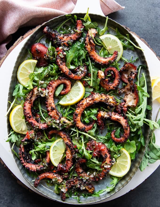 Large appetizer plate of grilled octopus recipe served over arugula, lemon sliced and drizzled with more olive oil marinade