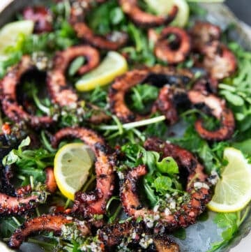 Close up of a plate of grilled octopus surrounded by lemon slices and arugula