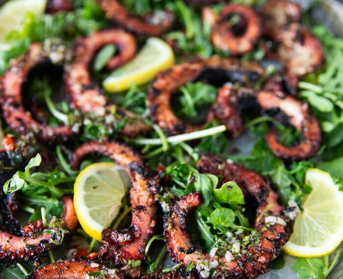 Close up of a plate of grilled octopus surrounded by lemon slices and arugula