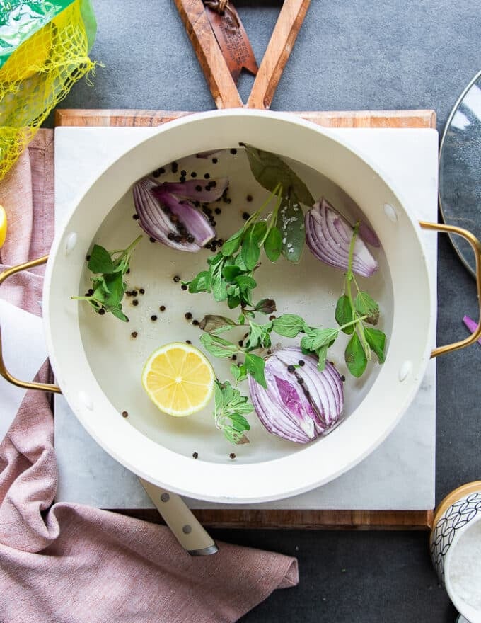 A pot filled with water, onion halves, lemon slices, fresh herbs, bay leaves, whole peppercorn and seasoning on the stove ready to boil for the octopus