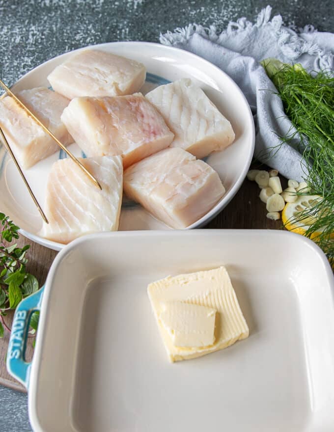 ingredients for halibut recipe including a plate with four halibut fillets, another plate with butter, lemon, herbs and lemon