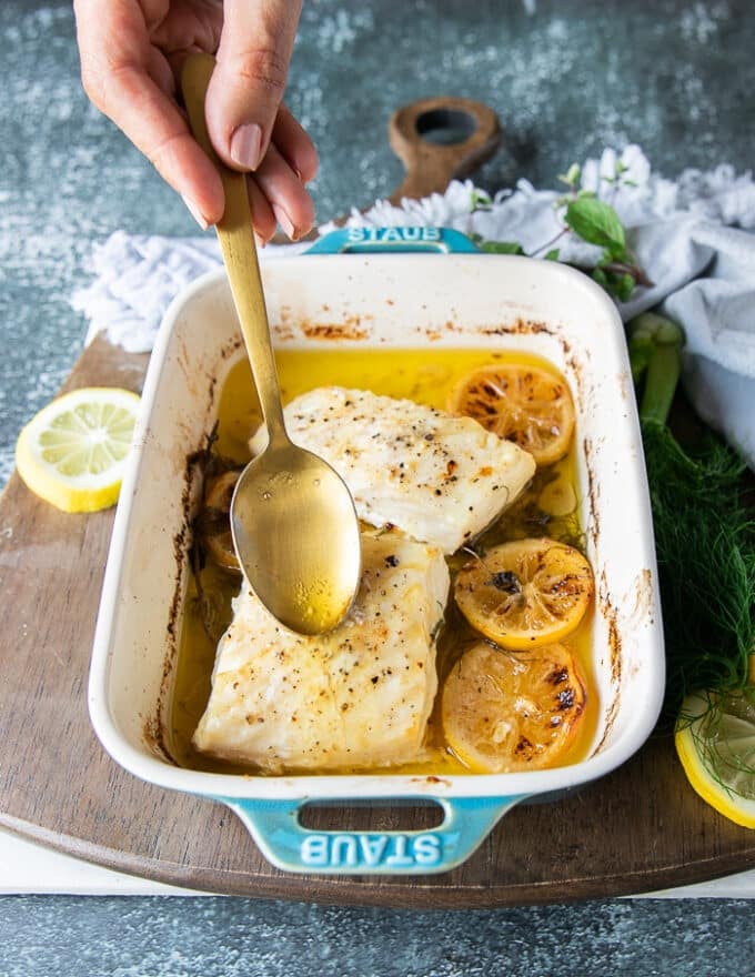 A hand scooping some of the butter from the bottom of the dish over the halibut to add even more butter flavor.