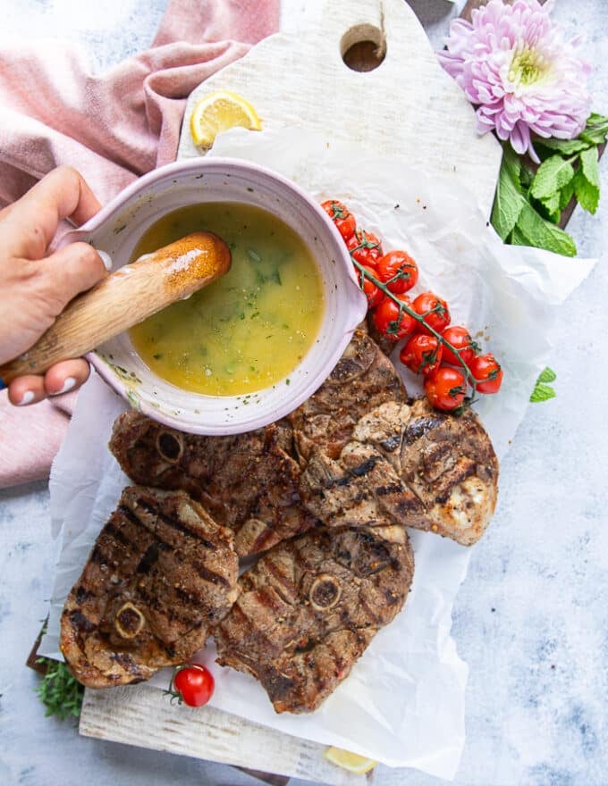 A hand pouring the herb butter over the grilled steaks
