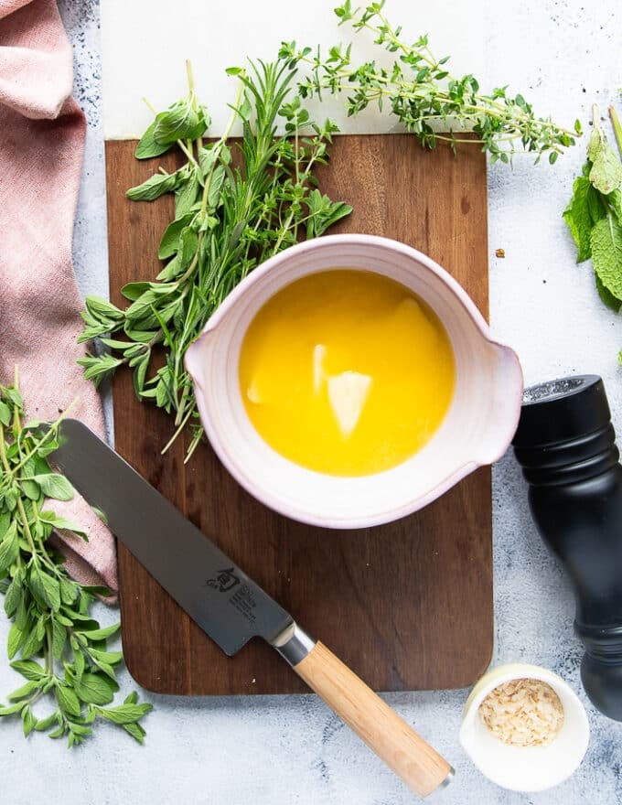 herb butter ingredients on a wooden board including melted butter in a bowl, some fresh herbs, salt and pepper, onions flakes