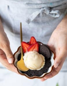 A hand holding an oreo mug cake close up with a scoop of ice cream and sliced strawberries on the side