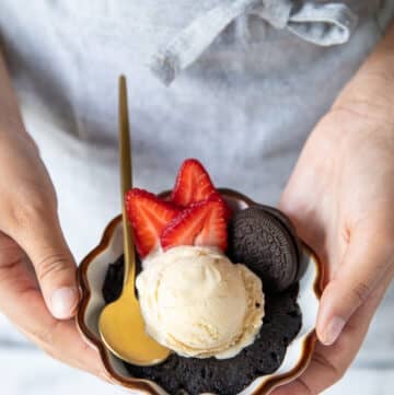 A hand holding an oreo mug cake close up with a scoop of ice cream and sliced strawberries on the side