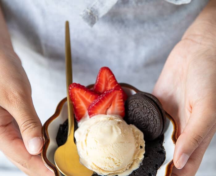 A hand holding an oreo mug cake close up with a scoop of ice cream and sliced strawberries on the side