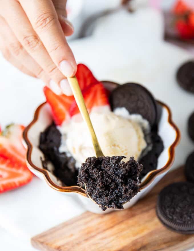 A hand taking a spoon of the oreo mug cake served with ice cream and showing close up the cakey texture of the cake!