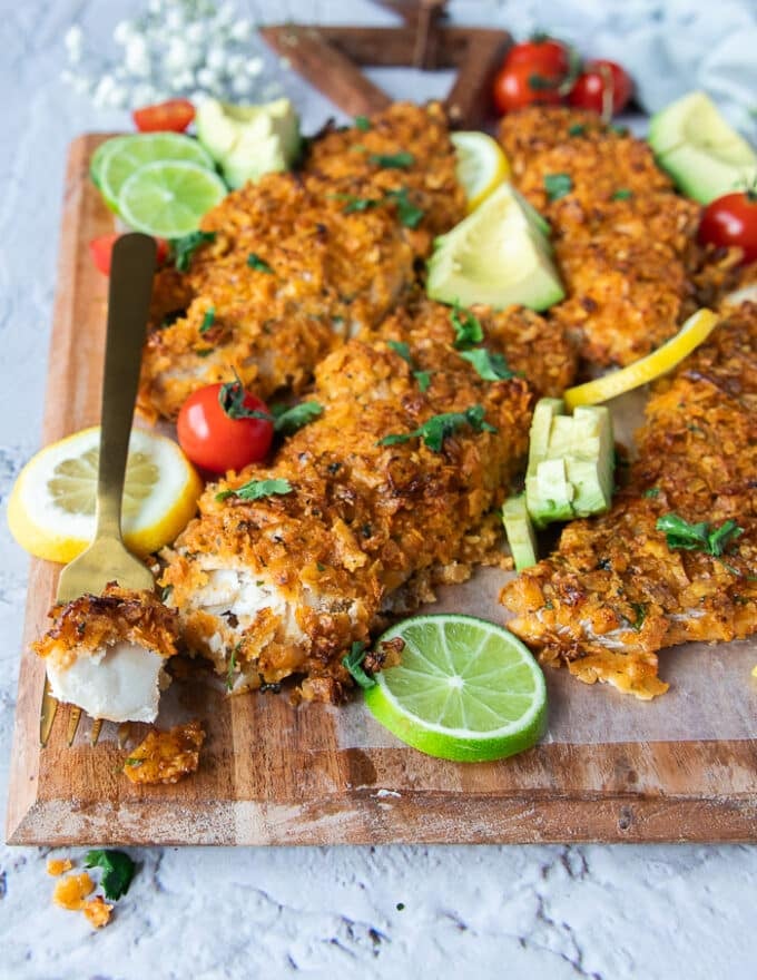 Baked haddock fish on a board with some cilantro, lime wedges and a fork cutting through the fillets