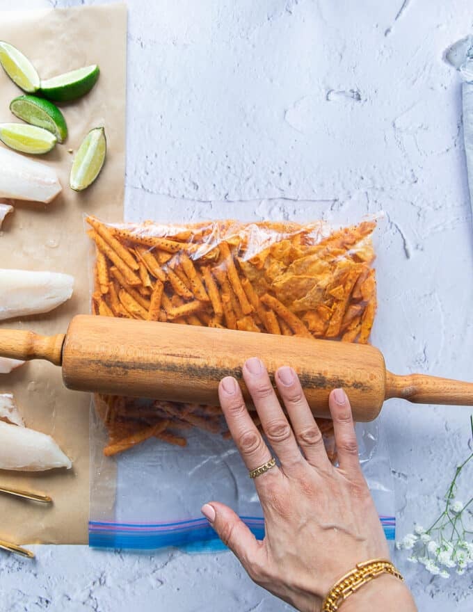 A hand holding a rolling pin and crushing some chips in a ziploc bag to make the crunchy coating