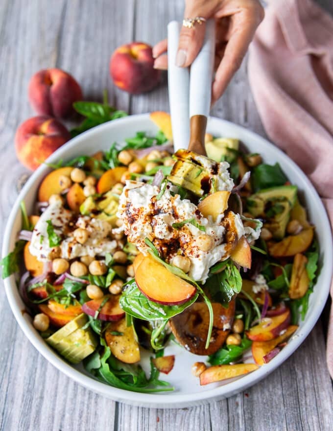 two hands serving some peach burrata salad with serving spoons showing close up of the burrata and peaches and balsamic dressing