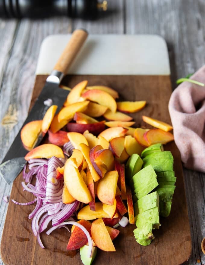 All ingredients for the peach burrata salad are sliced on a cutting board including sliced peaches, red onions, avocados