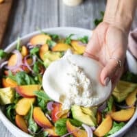 A hand holding the burrata cheese breaking it apart slightly over the peach salad