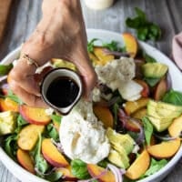 A hand pouring the balsamic syrup dressing over the assembled peach burrata salad