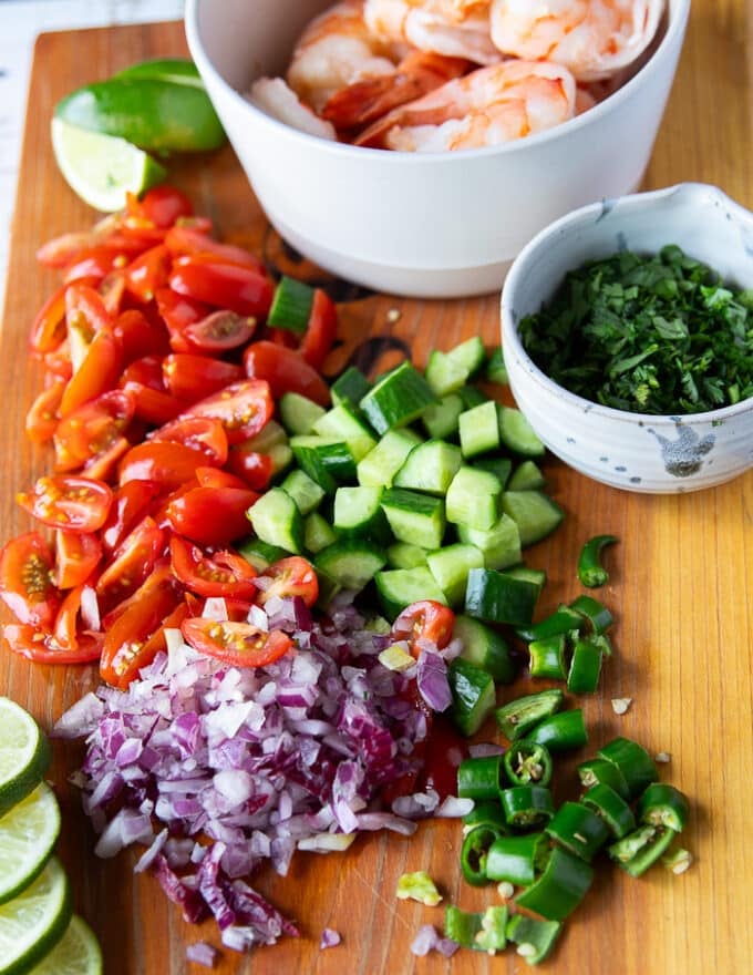 Ingredients for shrimp ceviche including diced onions, tomatoes, cucumbers, avocados, cilantro, the shrimp in a bowl, some diced jalapenos, lime juice