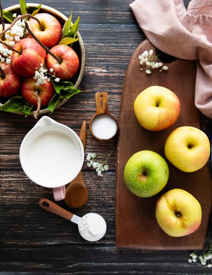 Apple crisp Ingredients including a variety of apples on a wooden board, a spoon of flour, a spoon of sugar, teaspoon of cinnamon , a cup of milk