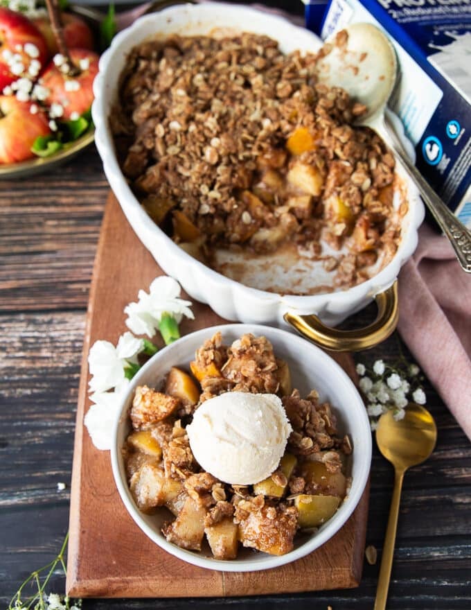 A small bowl of apple crispt topped with a scoop of vanilla ice cream near the large apple crisp plate and a spoon next to the plate