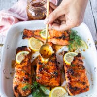 A hand pouring in some hot honey over the salmon right out of the oven garnished with lemon and dill