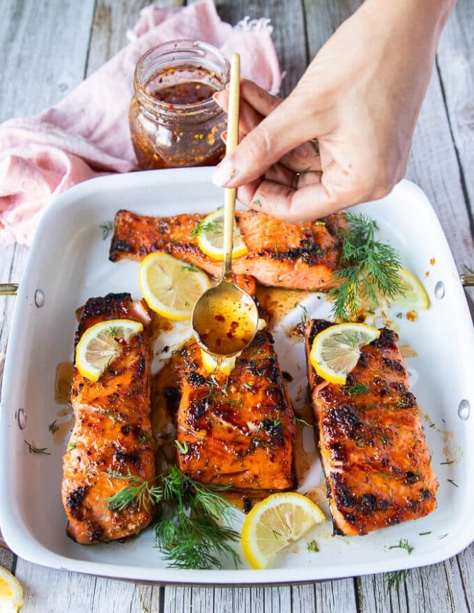 A hand pouring in some hot honey over the salmon right out of the oven garnished with lemon and dill 