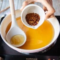 A hand adding in a small bowl of chilli flakes over the honey in a saucepan over medium heat