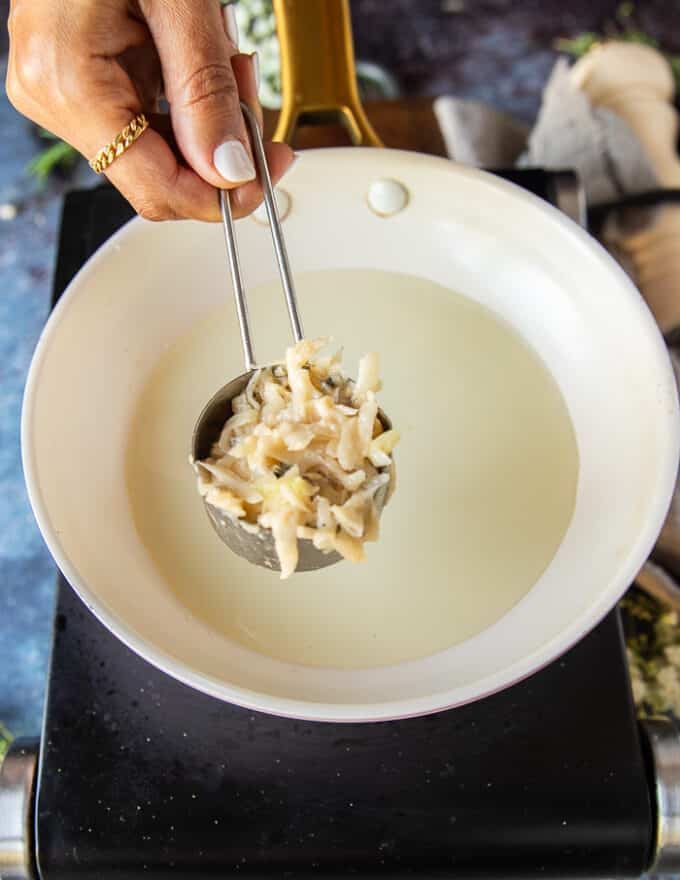 A hand holding a quarter cup measuring cup filled with potato pancake mixture portioned and ready to cook it in hot oil. 