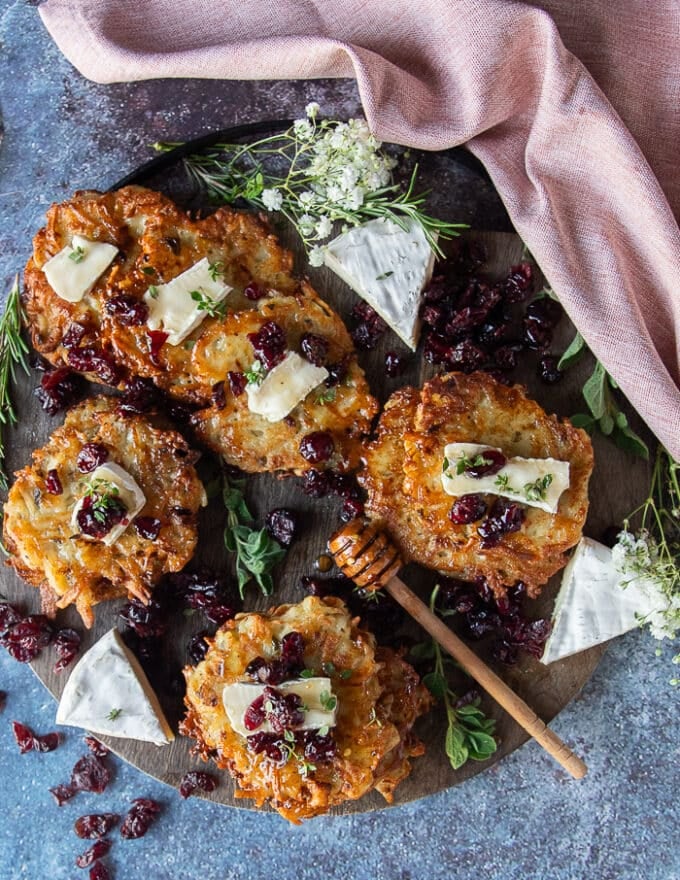 A board filled with stacks of potato pancakes topped with cranberries and brie cheese, surrounded with a honey jar
