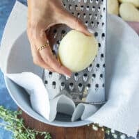 A hand grating the peeled potatoes using a box grater over a bowl lined with kitchen towel to hold excess moisture