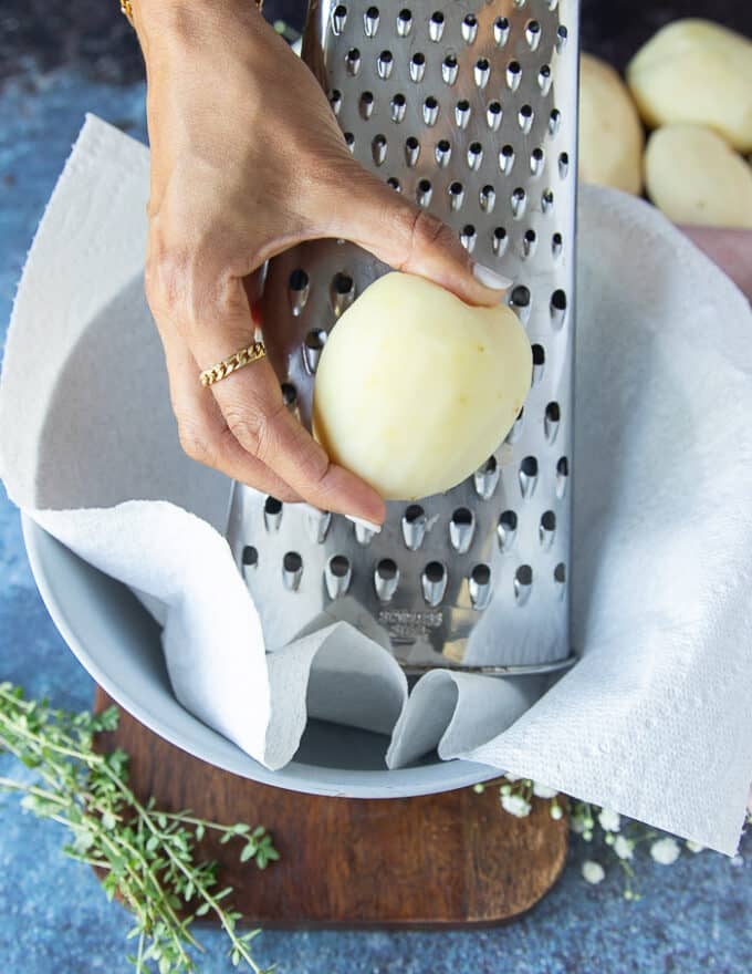 A hand grating the peeled potatoes using a box grater over a bowl lined with kitchen towel to hold excess moisture