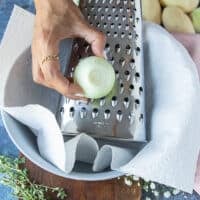 A hand holding onions and grating them using the same box grater over the bowl of grated potatoes lined with kitchen towel
