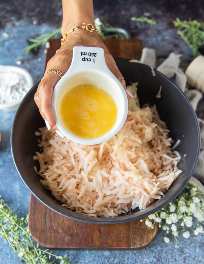 A hand holding an egg and pouring it over the squeezed potatoes and onions in a new bowl