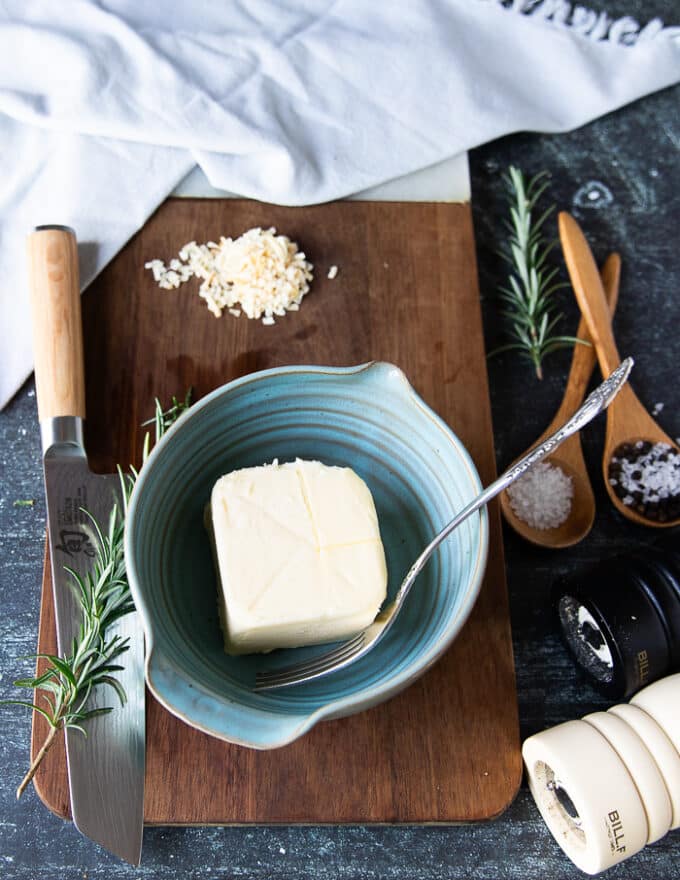 ingredients for rosemary butter on a board including butter in a bowl, fresh rosemary leaves, some onion flakes, salt and pepper and block of parmesan cheese
